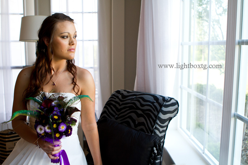 bride gazes out of window holding a bouquet of purple flowers and peacock